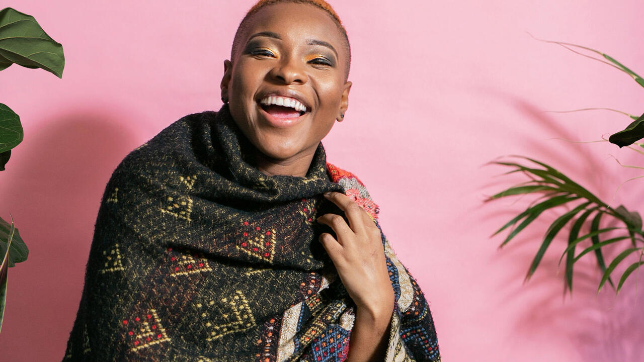A young woman with dark skin is smiling towards the camera with her eyes squinted. She is against a pink background with plants decorating it