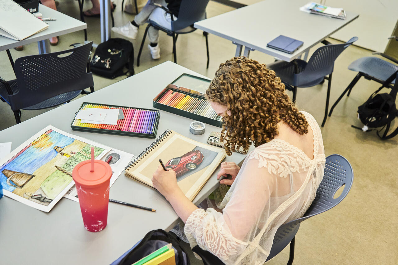 Student works at a table on colorful drawings in colored pencil with two colored pencil sets on the table.