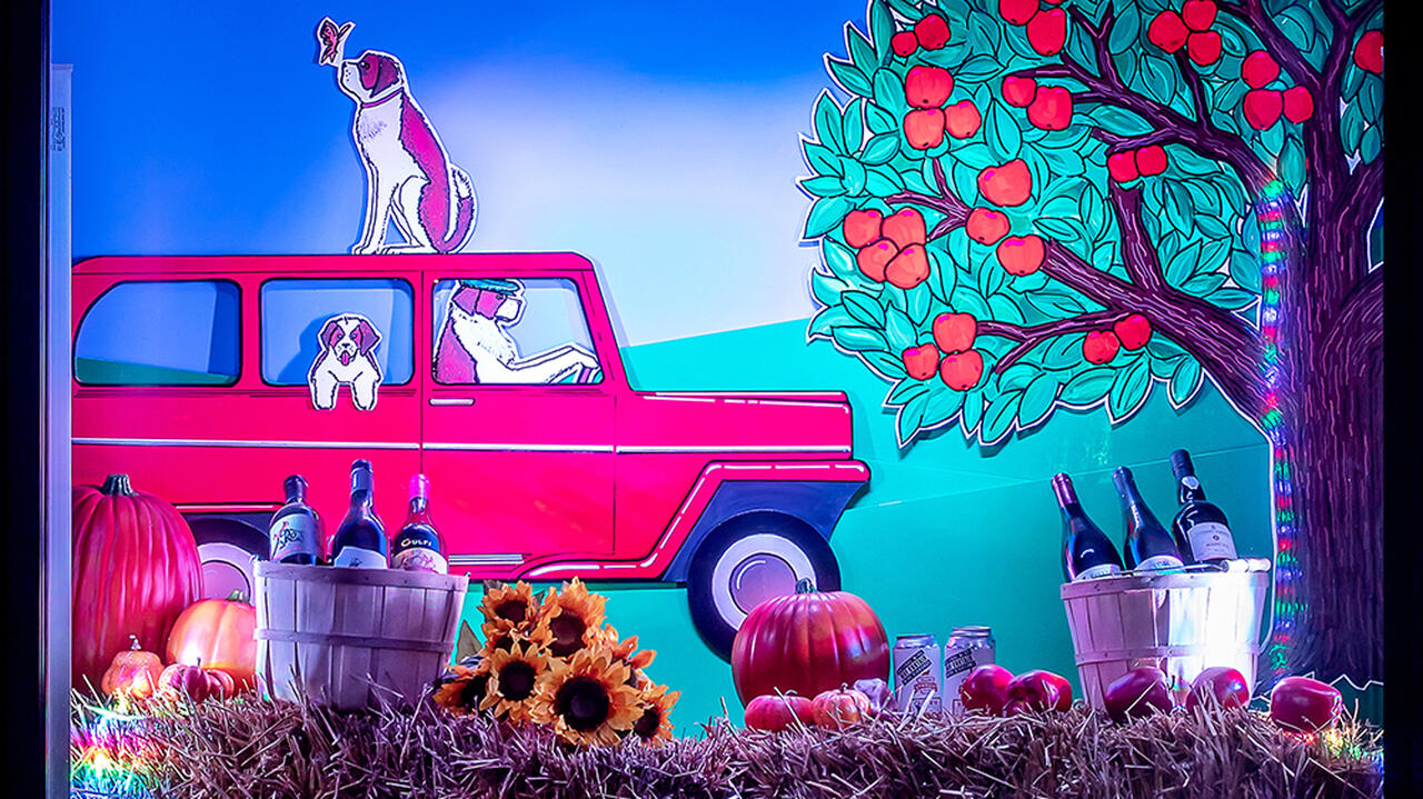 A display featuring a red car in the back with three dogs inside, to the right of the car there is an apple tree. In the front of the display you can see alcohol, sunflowers and pumpkins