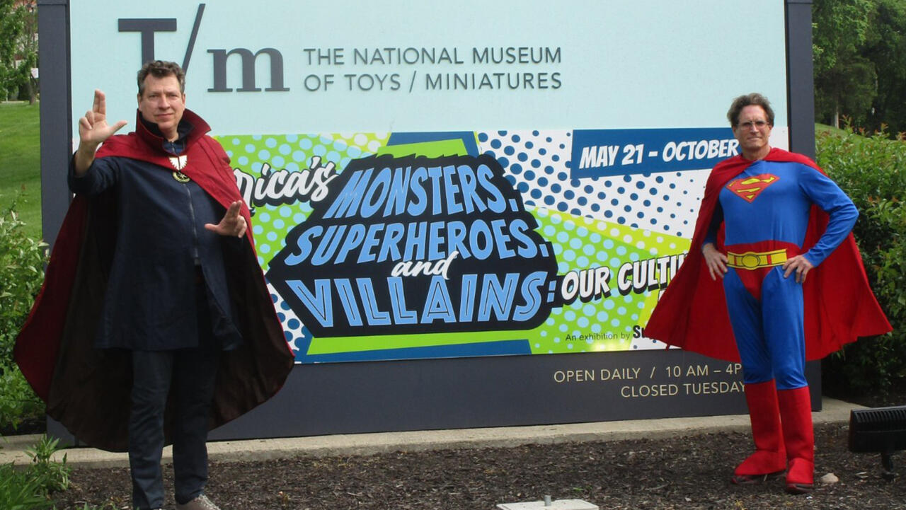 Two men stand outside infront of a sign that reads "MONSTERS SUPERHEROS AND VILLAINS" They are both dressed up respectively as Dr.Strange and Superman