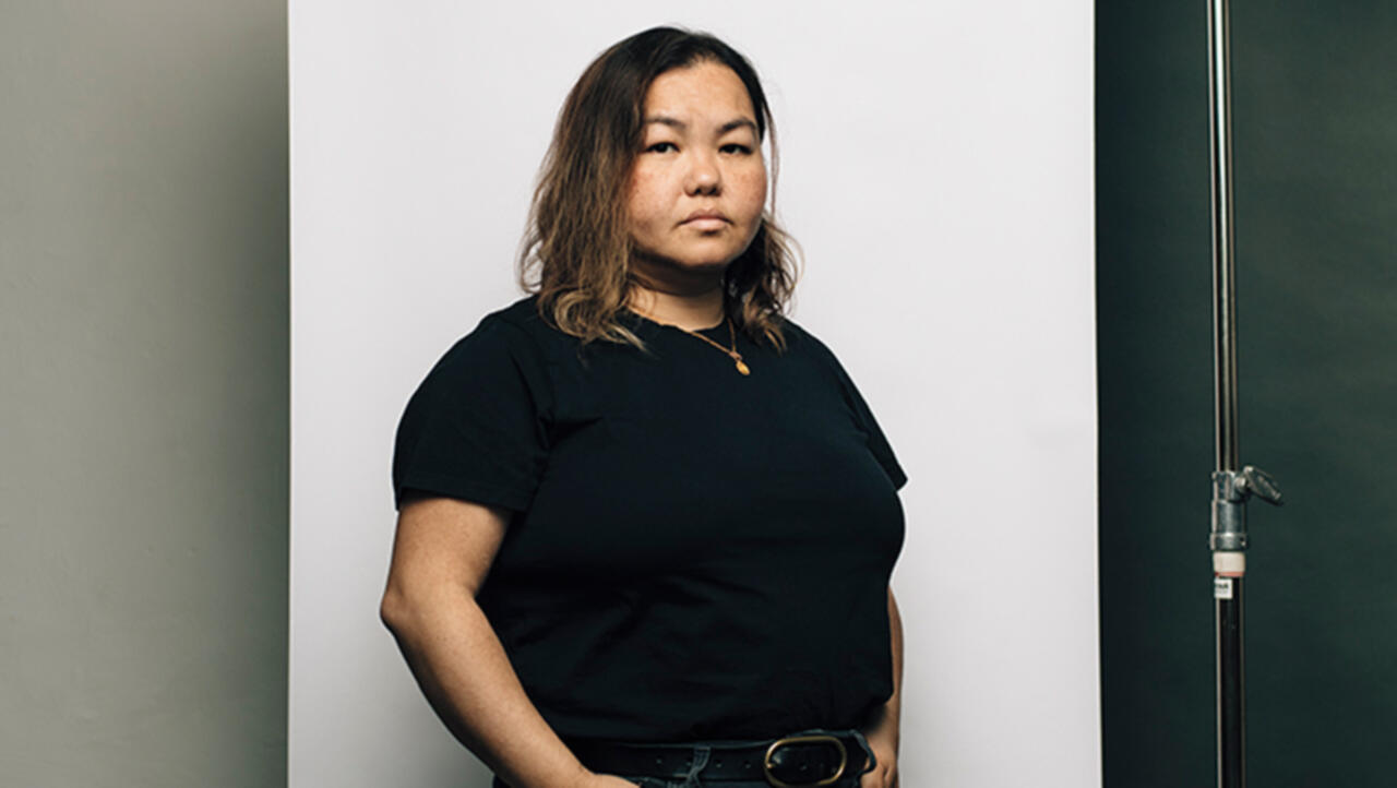 A woman in a black shirt stands against a white backdrop, her expression is flat and staring directly into the camera with one hand popped out on her hip