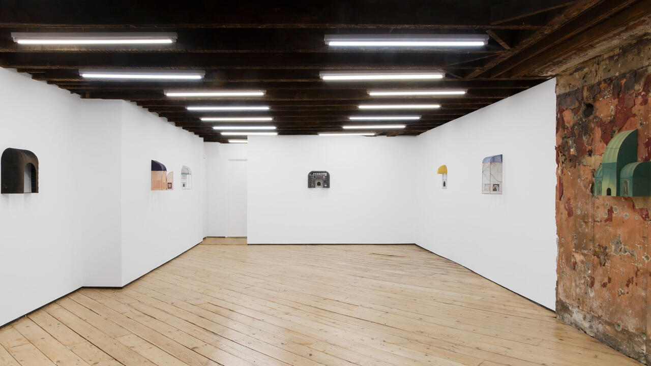 An interior of a room. The room has white walls, fluorescent lighting and a wooden floor.  Displayed on the walls are different pieces that are sculptures resembling window awnings