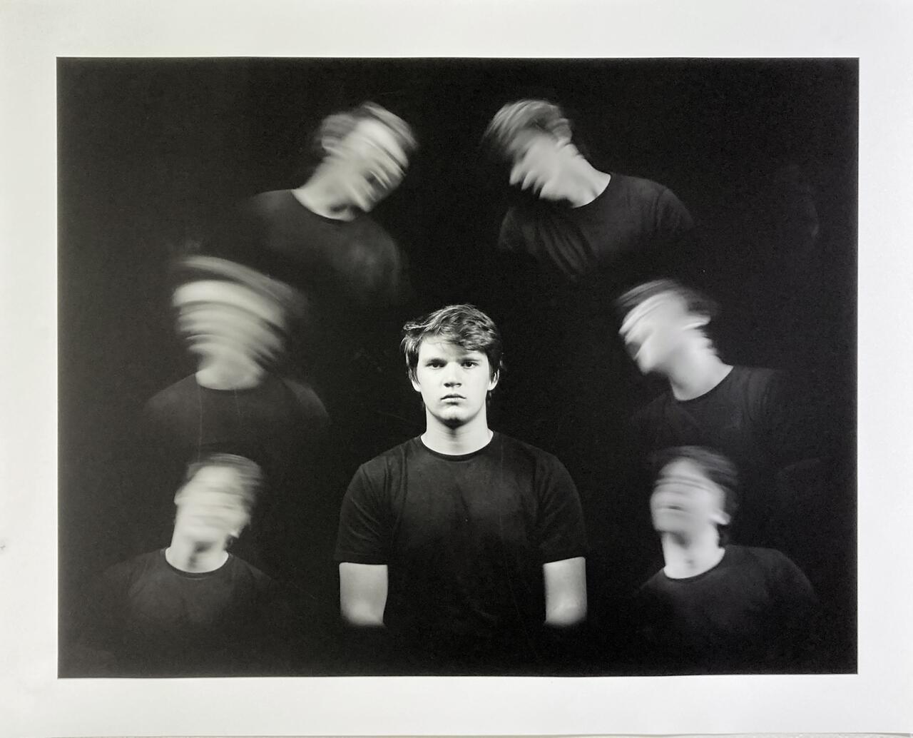 Black and white photograph featuring one student in several poses from the torso up. Central is the student looking directly into the camera. On each side of the central figure are three images of the student with a blurred head showing different movement and facial expressions.