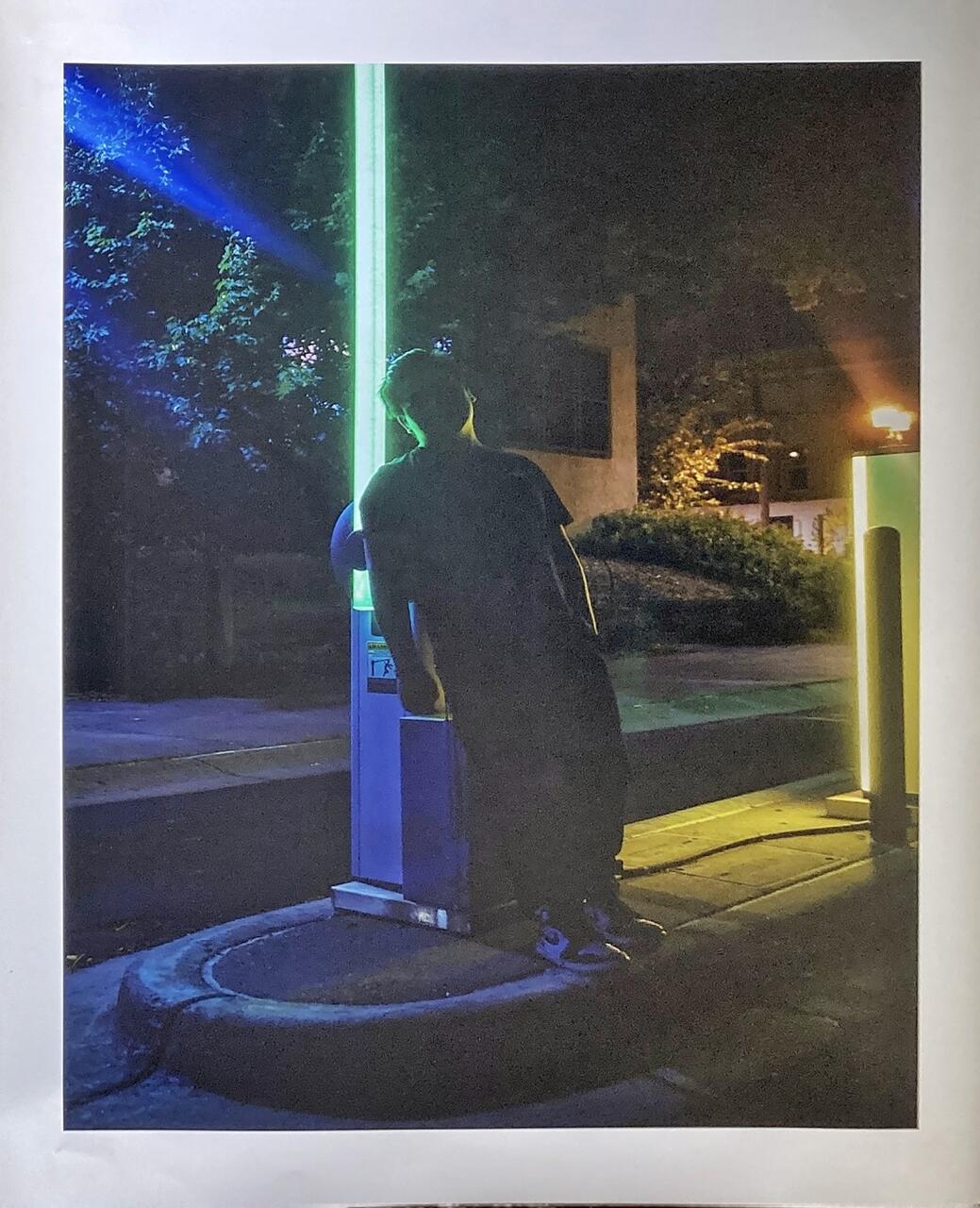 Color photograph of student standing outside at night, leaning on a parking gate arm that is lit up in green. Dimly lit trees and other parking structures can be seen in the background.