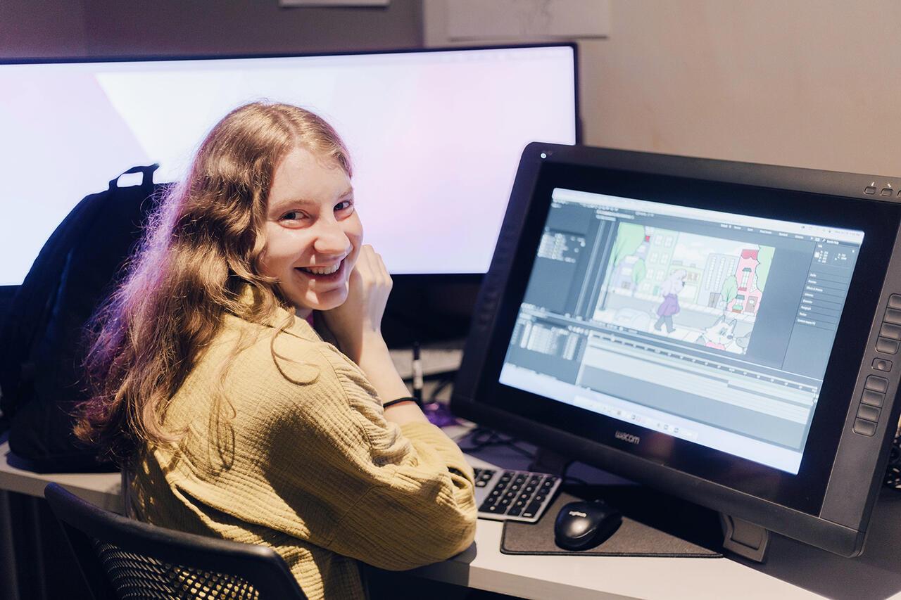 Student sits at an animation computer with an animation still showing on her computer. Student looks over shoulder at camera and smiles. Her backpack can be seen in the background.