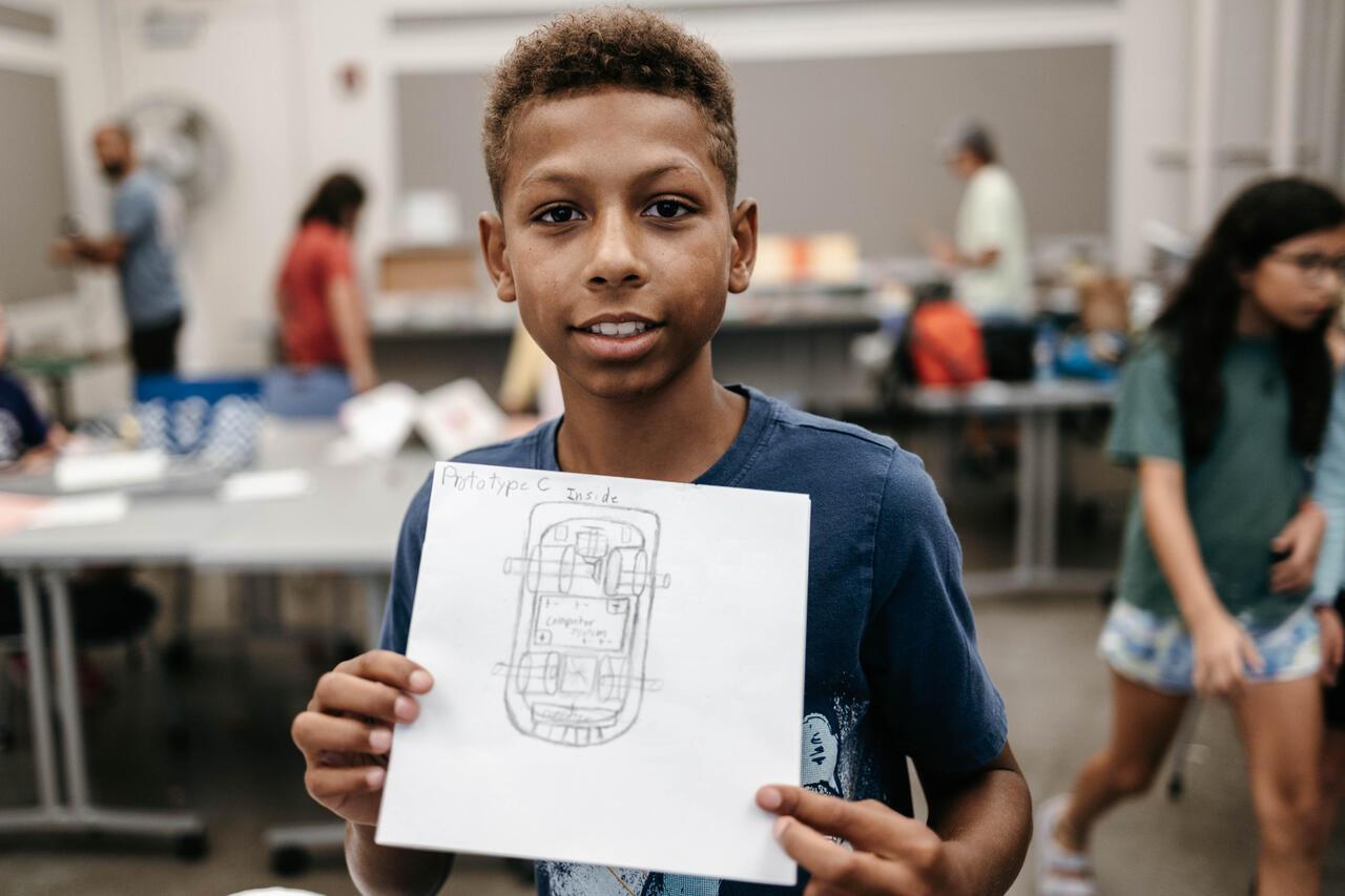 Student holds up pencil drawing of a prototype and stands in front of an active classroom.