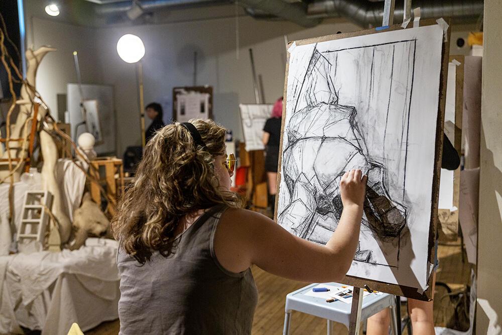 Teen student sits at drawing board working on a charcoal still life. Still life and other students can be seen in the background.