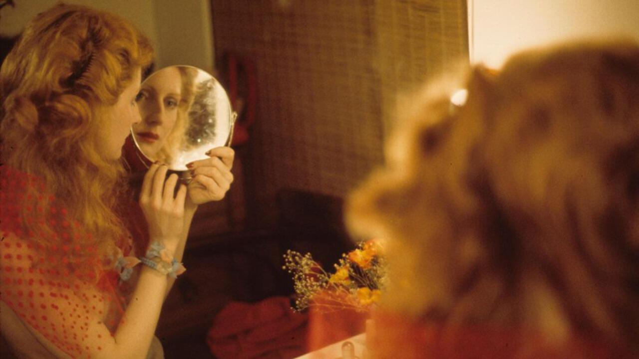 A screenshot from "The Disappearance of Shere Hite" it shows a woman looking into a hand mirror reflecting in a vanity