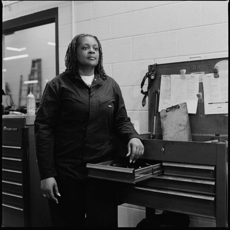 A black and grey portrait of ATU worker Kira Heywood standing infront of their open toolbox. She's looking off to the right.