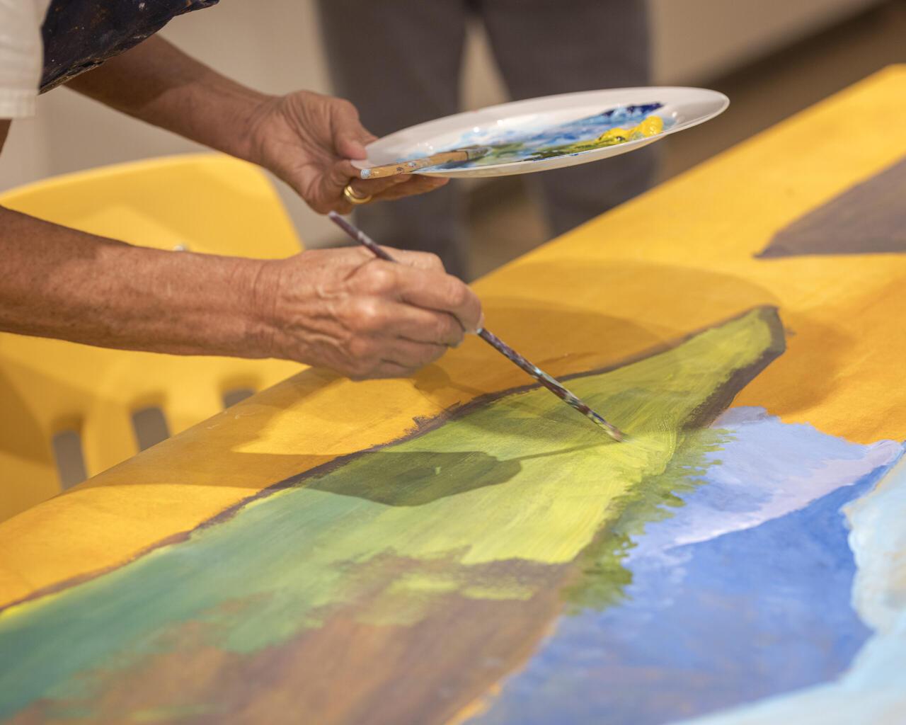 A pair of hands holding a paint pallete in the left, and a brush on the right. They're applying paint to a green portions of a multi-color canvas.