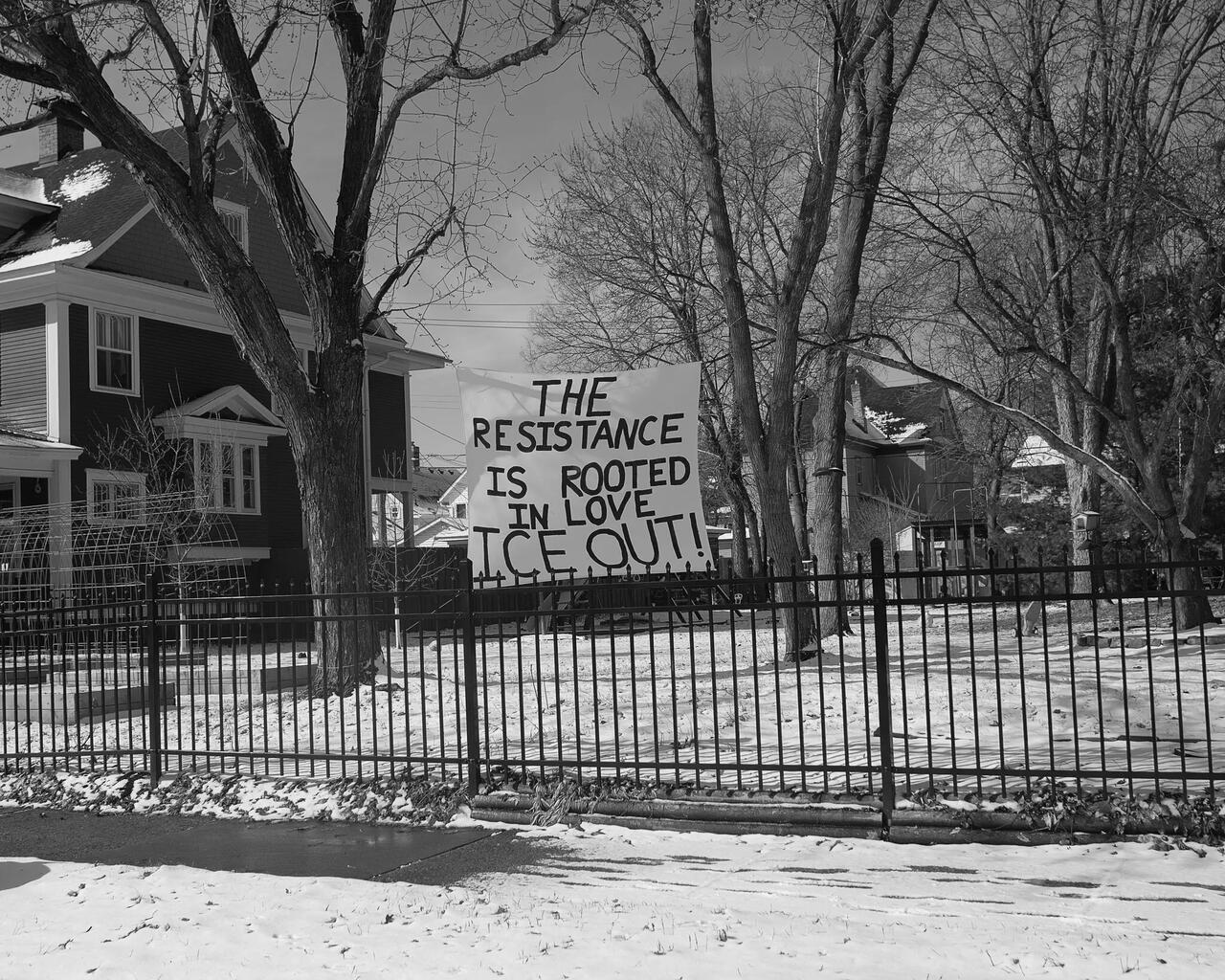 A photograph of a handwritten sign on a fence that reads: “The resistance is rooted in love. ICE out!” 