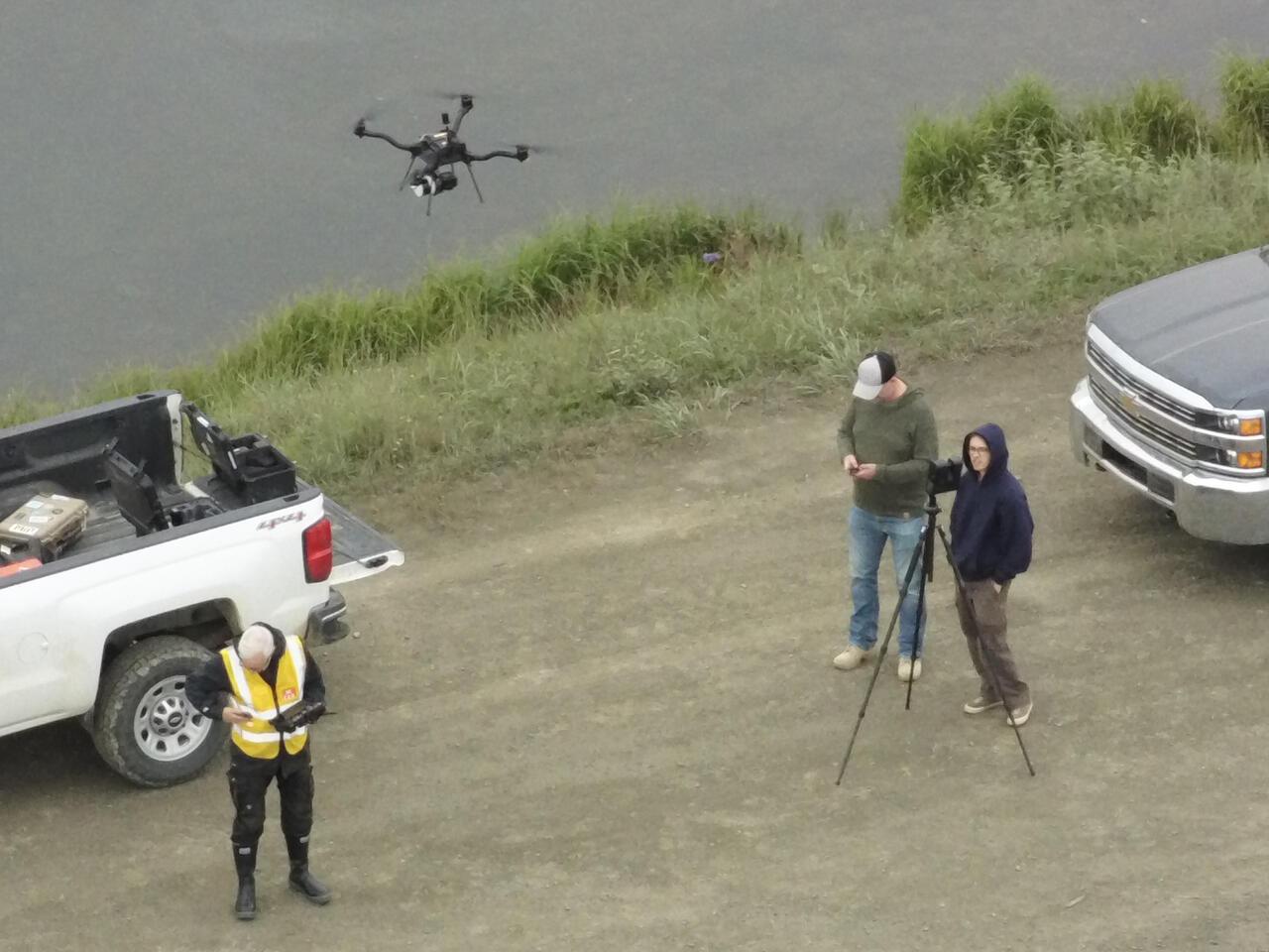 Three people stand in a field with a drone hovering overhead