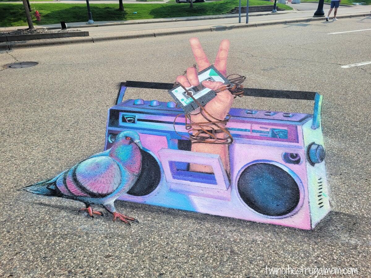 Street chalk art of a pastel purple and blue boom box. A hand is popping out of the place where you would inseart a tape, making a peace sign and holding a mix tape with wires wrapped around it. A pigeon is standing on the left side of the boom box.