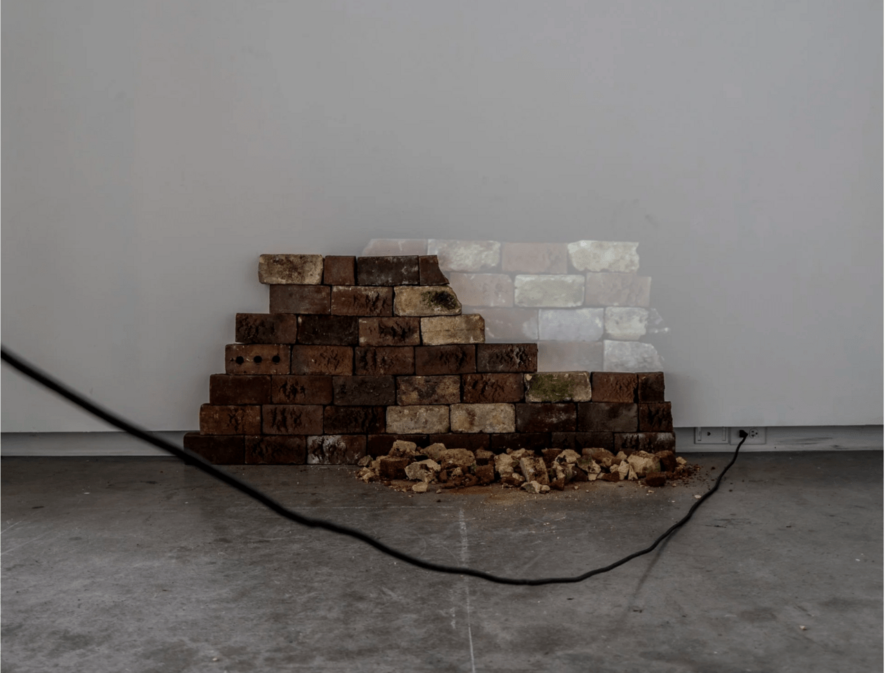 A photo of a sculpture titled Ones Dismantling Impedes the Others by Evan McHenry. The sculptures is a stakc of bricks stacked, with a video projection playing the bricks falling down.