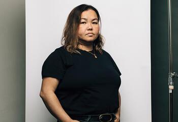 A woman in a black shirt stands against a white backdrop, her expression is flat and staring directly into the camera with one hand popped out on her hip